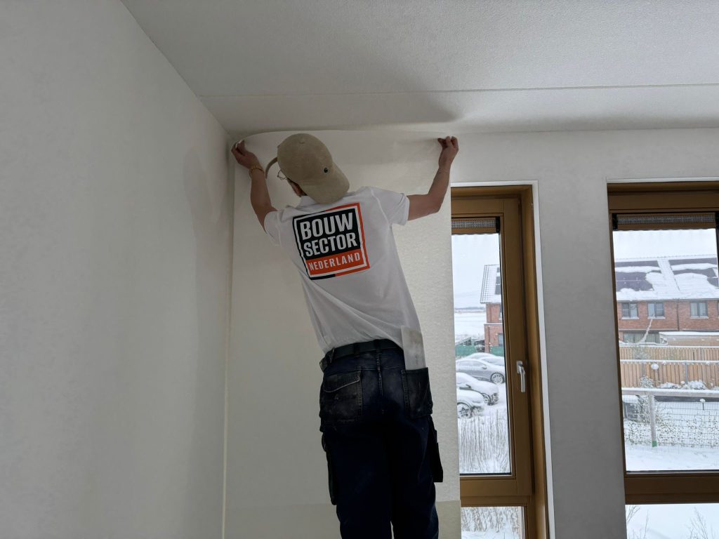 Behanger van Bouwsector Nederland drukt een baan renovlies tegen de bovenkant van een witte wand, naast twee ramen met houten kozijnen