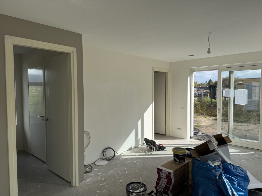Woonkamer van een net opgeleverde woning tijdens de afwerking, met verfemmers, dozen en kabels op de kale vloer
