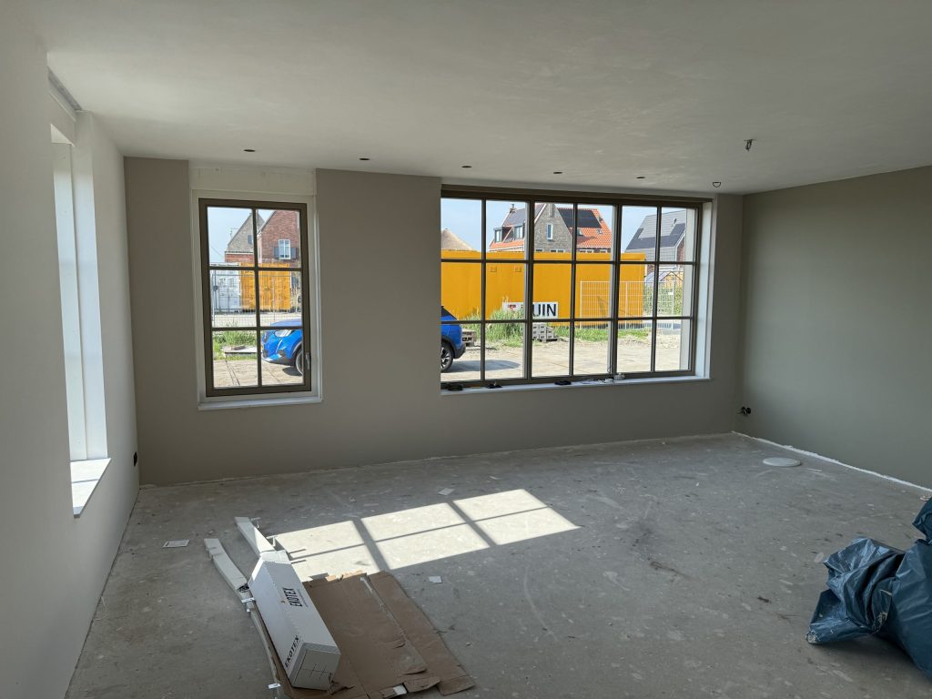 Kamer in een nieuwbouwwoning met grijsgroene wanden, een kale vloer en een groot raam met roeden
